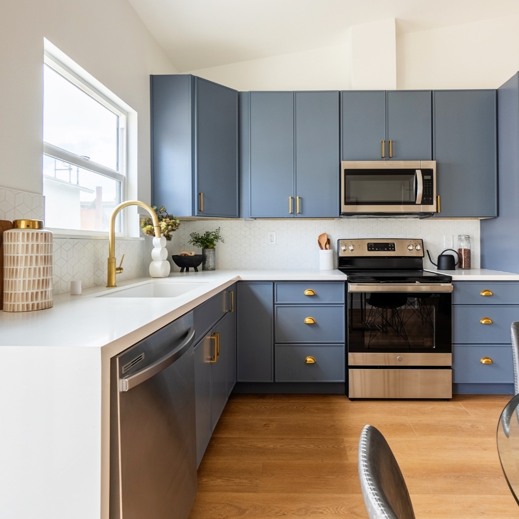 A modern kitchen in an ADU featuring elite finishes. The space includes sleek blue cabinetry with gold hardware, complemented by stainless steel appliances. The countertops are a pristine white, paired with a stylish backsplash featuring geometric patterns. A large window above the sink allows natural light to flood the space, highlighting the elegant gold faucet. The kitchen exudes contemporary sophistication with its clean lines, high-quality materials, and thoughtful design details.