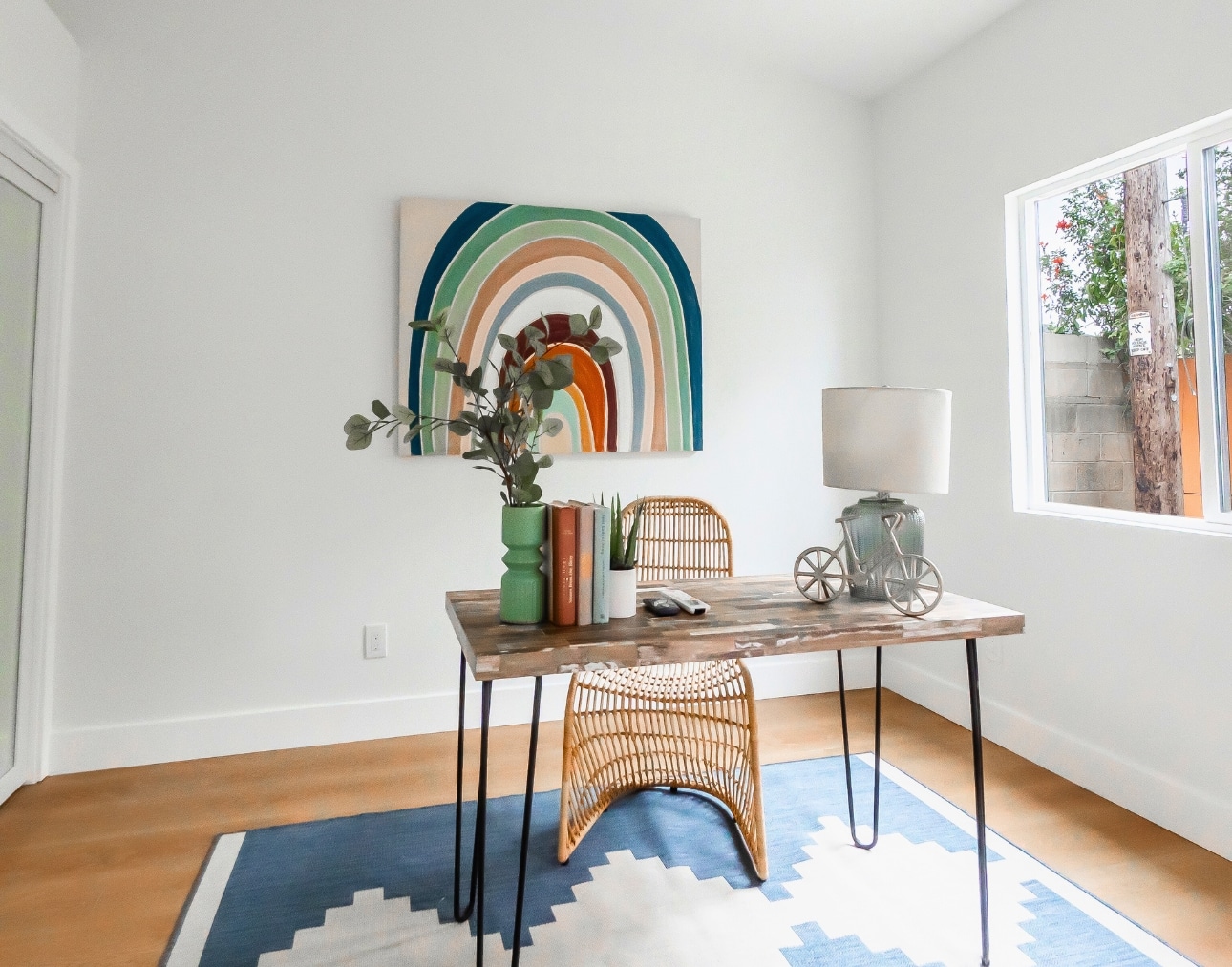 A modern ADU home office with natural light, rattan furniture, a rustic wood desk, and a colorful rainbow wall art, showcasing the benefits of ADUs for functional workspace solutions.