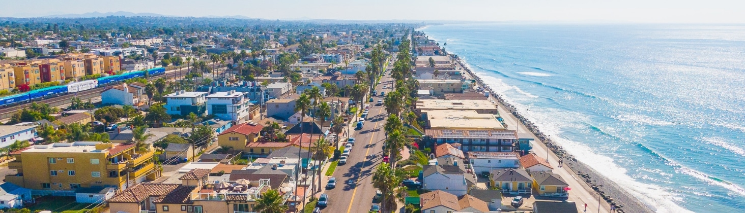 Aerial view of a coastal city featuring a long beachfront, houses, buildings, and a main road running parallel to the ocean. The sea is calm with gentle waves hitting the shore.