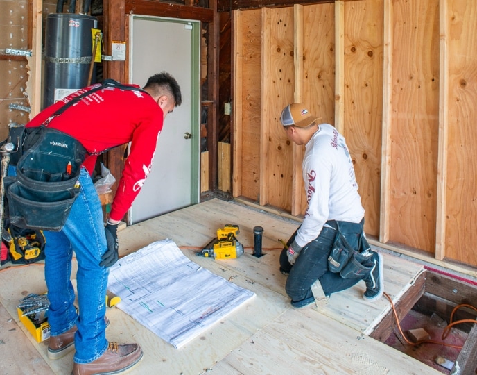 Two builders reviewing blueprints inside a partially constructed space, focusing on layout and structural planning. Highlights the construction and space requirements for pool houses and ADUs, where ADUs often require more extensive planning for utilities and living amenities compared to the simpler recreational purpose of pool houses.