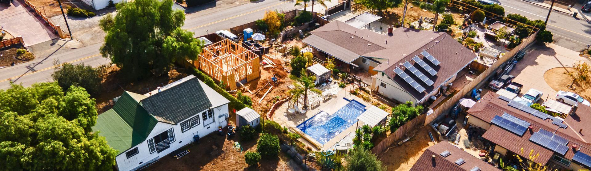 An aerial view of a residential property with an ADU under construction next to existing homes with solar panels and a pool. Caption: Adding an ADU increases property value, but working with an experienced contractor can minimize tax implications and ensure seamless integration with your property.