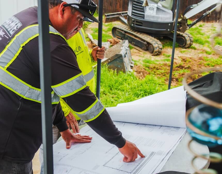 Construction workers reviewing ADU blueprints on-site with excavation equipment in the background.