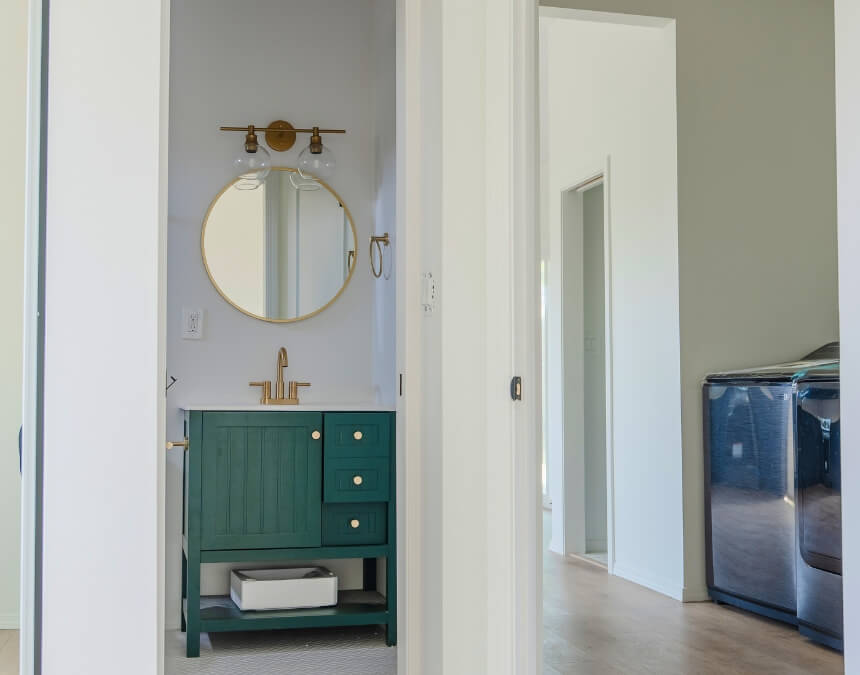 Compact bathroom with green vanity, round mirror, and gold fixtures in new ADU
