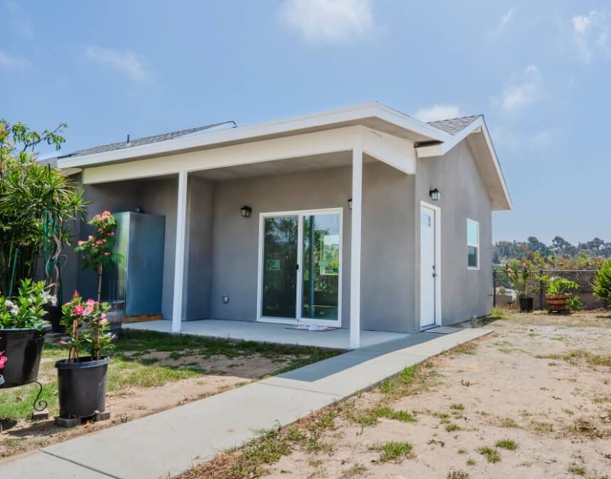 Newly constructed detached ADU with gray stucco exterior, white trim, a covered front porch, and surrounding dirt landscaping.