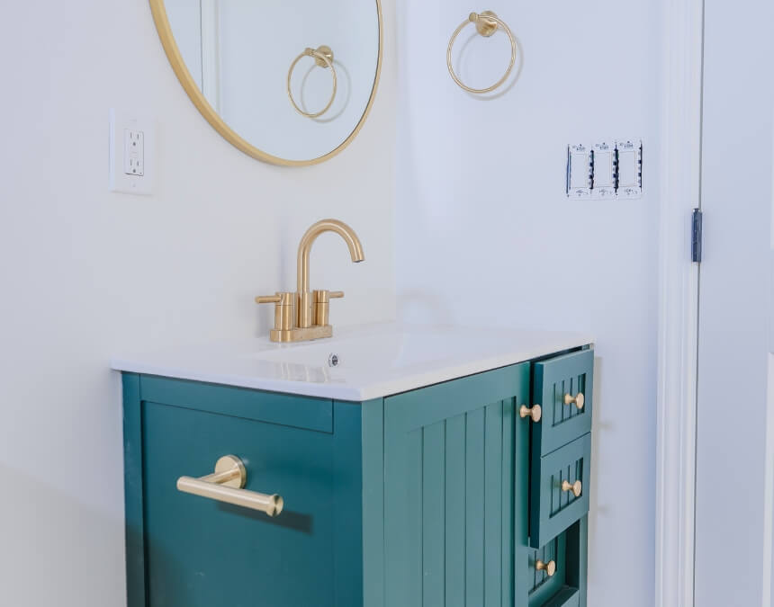 Modern bathroom vanity with brass fixtures, teal cabinetry, and round mirror on a white wall.