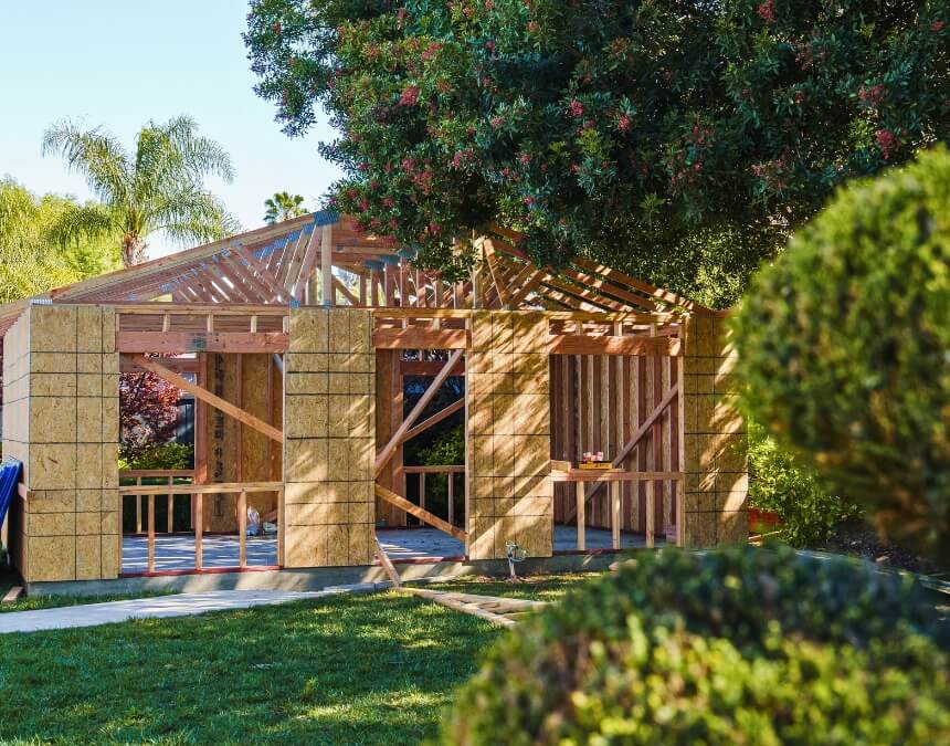 Framed structure of an ADU mid-construction, partially hidden behind trees and surrounded by a well-maintained lawn.