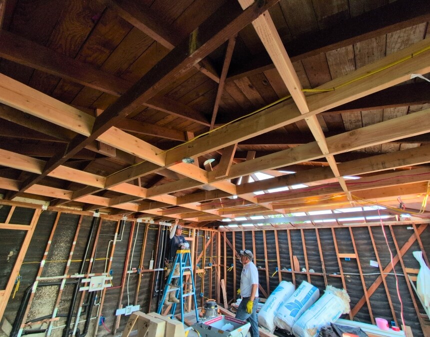 Inside view of ADU rough framing with visible wiring, plumbing, and two workers inspecting utility installations