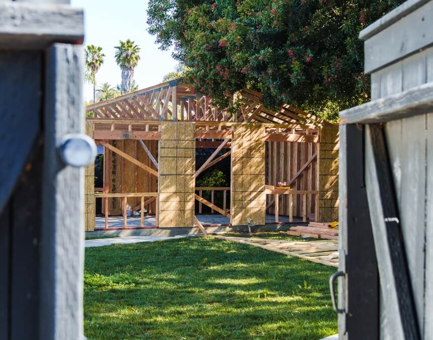 Wooden ADU framing in progress behind a fence, partially concealed by surrounding trees and structures.