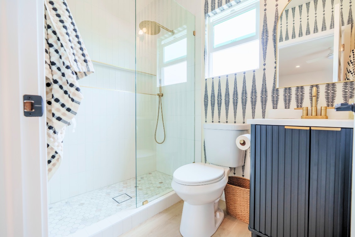 Compact ADU bathroom with patterned wallpaper, glass shower, and navy vanity with gold hardware