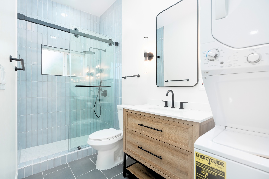 Small ADU bathroom with wood vanity, glass shower, and in-unit stacked washer and dryer